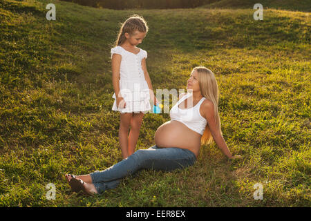 Cute little girl watering her mother's pregnant belly in summer nature. Big sister wants her brother to grow faster. Stock Photo