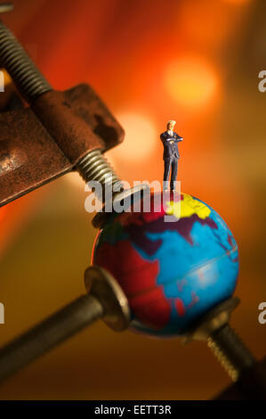 Conceptual macro shot of a small figurine of businessman standing on top of a globe that is being squeezed by a clamp Stock Photo