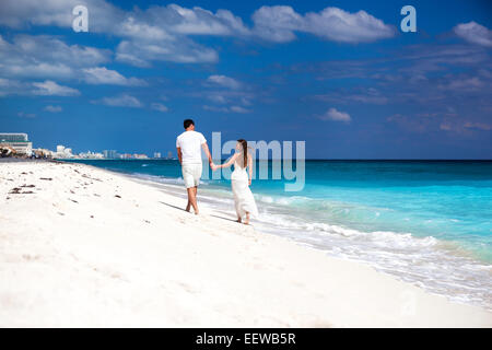 Young beautiful newlyweds in love holding hands and walking away on tropical sandy beach, back view Stock Photo