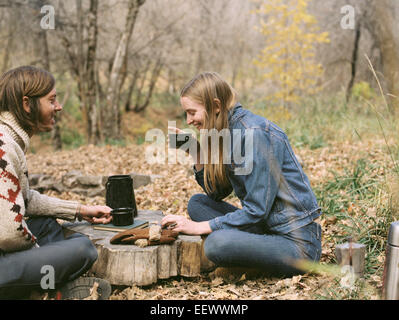 Smiling couple sitting on the ground in a forest, drinking coffee. Stock Photo