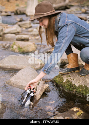 Young blond woman wearing a hat, putting drink bottles in a river. Stock Photo