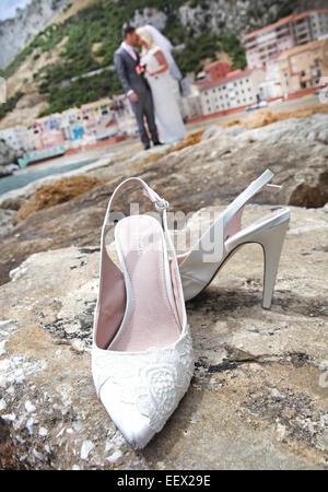 Bride and groom kissing on stone breakwater in Gibraltar with Catalan Bay village in  background and wedding shoes in foreground Stock Photo
