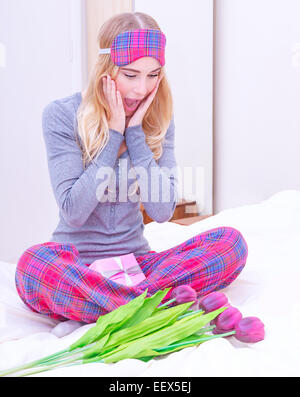 Beautiful woman sitting on the bed and enjoying fresh tulip bouquet and gift,  surprise expression, morning in Valentine day Stock Photo