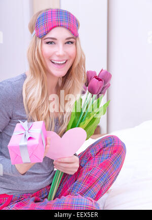 Portrait of happy smiling girl sitting on the bed in pajama and enjoying romantic gift in Valentine day, receive flowers Stock Photo