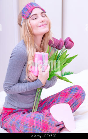 Happy woman sitting on the bed wearing cute pajama and with closed eyes with pleasure enjoying flowers and romantic gift Stock Photo