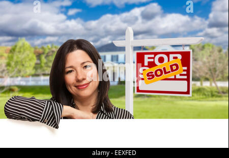 Pretty Hispanic Woman Leaning on White in Front of Beautiful House and Sold For Sale Real Estate Sign. Stock Photo