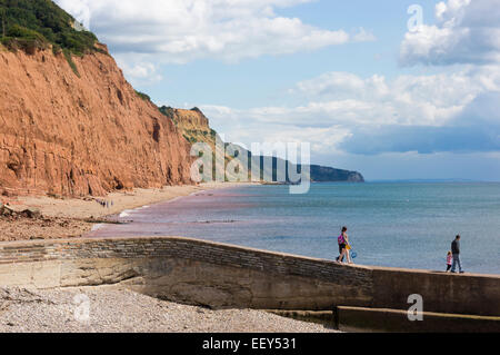 Red Cliffs at Sidmouth, East Devon, England, UK - with family walking along a stone jetty in summer Stock Photo