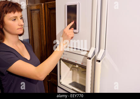 Young woman operating touch pad on web enabled refrigerator internet of things appliance in kitchen Stock Photo