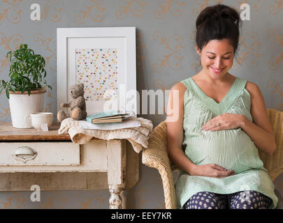 Pregnant woman sitting in chair touching abdomen smiling Stock Photo