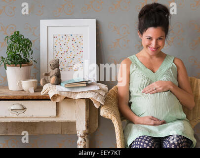 Pregnant woman sitting in chair touching abdomen smiling Stock Photo