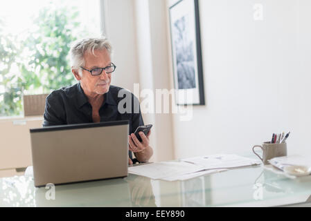 Senior man sitting in home office, using laptop and cell phone Stock Photo