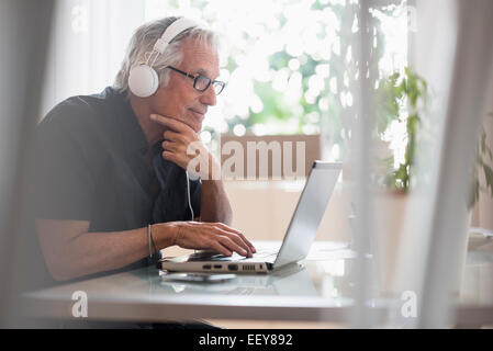 Senior man sitting in home office wearing headphones and using laptop Stock Photo