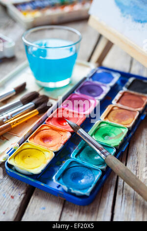 Set of watercolor paints,  art brushes,  glass of water and easel with painting on vintage wooden background. Stock Photo