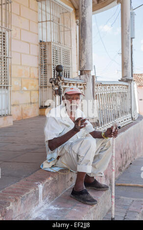 Friendly but infirm local old man dressed in white clothing with a walking stick sitting on steps smoking a Cuban cigar in downtown Trinidad, Cuba Stock Photo
