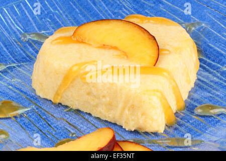 Heart shaped peach bavarian cream dessert (bavarese) with peach sauce and peach slices on a transparent glass plate over a blue Stock Photo