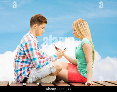 couple with smartphones sitting on bench over sky Stock Photo