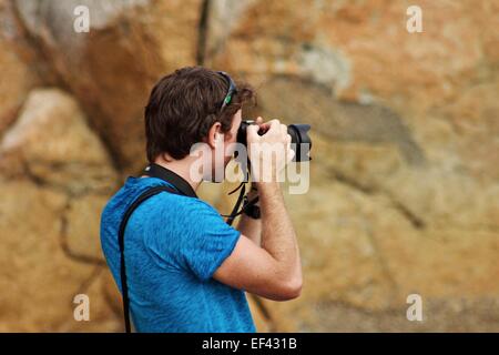 Young male photographer taking photos with DSLR camera outdoors near rocky formation. Stock Photo
