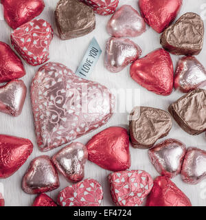 many heart shaped chocolates in colorful wrapping spread out over a table. The largest of which has a tag that says I love you. Stock Photo