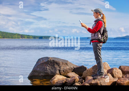 Young woman tourist with map and backpack standing on lake shore. Big rocks and wide lake on background. Stock Photo