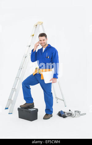 Smiling repairman with toolbox and ladder using cellphone Stock Photo