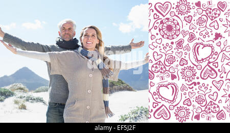 Composite image of carefree couple standing on the beach in warm clothing Stock Photo