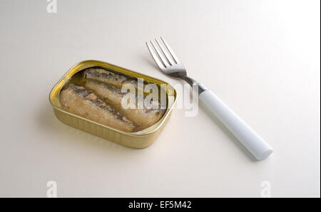 An opened can of sardines in oil and water with a fork to the side of the tin on a white table top illuminated by window light. Stock Photo