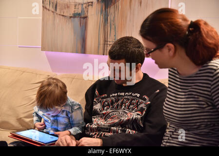 Two adults watching a young boy (2 1/2 yrs) sat on the sofa playing with an iPad Stock Photo