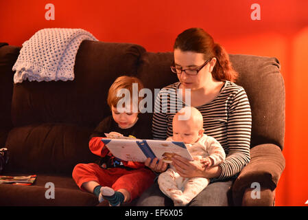 A mother sat on a sofa reading to her two children, one of five months and the other two and a half years Stock Photo