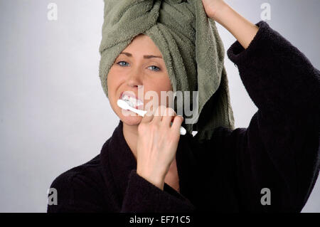 Young woman, age 20-25, brushing her teeth, holding green towel on her head, wearing black bath robe. Stock Photo