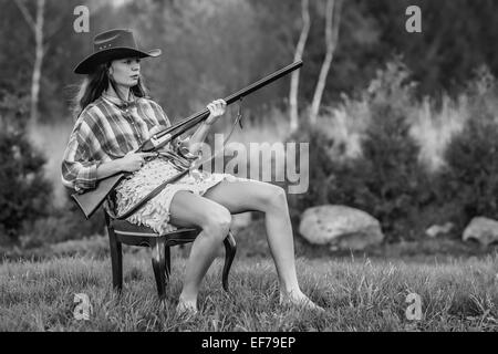 Black and white photography of a girl in cowboy hat sitting in a fancy chair with a shotgun in a backyard. Stock Photo