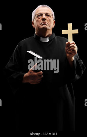 Vertical shot of a mature priest holding a cross and looking up on black background Stock Photo