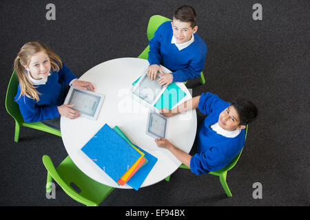 Overhead view of boys and girl sitting at round table with digital tablets, looking at camera Stock Photo