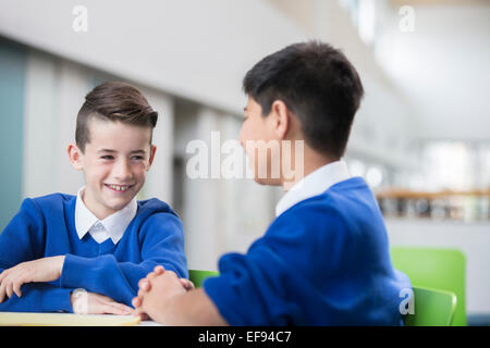 Two smiling schoolboys wearing school uniforms talking at desk in classroom Stock Photo