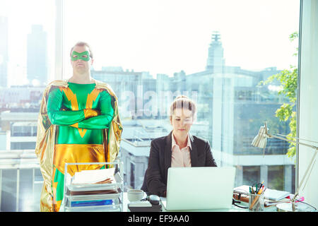 Superhero standing near businesswoman working in office Stock Photo