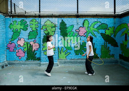 2 school girls skipping in the playground on the roof at their inner city Primary School with graffiti on the wall behind Stock Photo