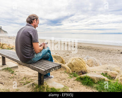 Man sitting on a bench texting on his cell phone at Arroyo Burro Beach in Santa Barbara, California. Stock Photo