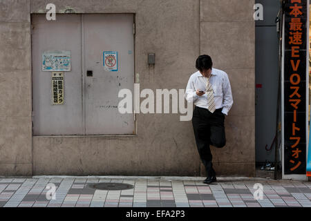 A Japanese salaryman or male office worker leans against a wall as he checks his cellphone in the street in Shinjuku, Tokyo, Japan. Stock Photo