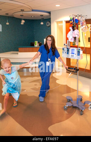 A smiling child life specialist at CHOC Children's Hospital in Orange, CA, plays with a young girl chemotherapy cancer patient in the hospital lobby. Note bald head and intravenous 'tree' and computer. Child life specialists support patients and families in the hospital environment. Stock Photo