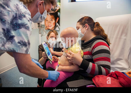 A child life specialist at CHOC Children's Hospital in Orange, CA, holds a computer tablet program to distract a young Hispanic girl patient undergoing a port access procedure for chemotherapy. Note masks, nurse, and bald head. Stock Photo