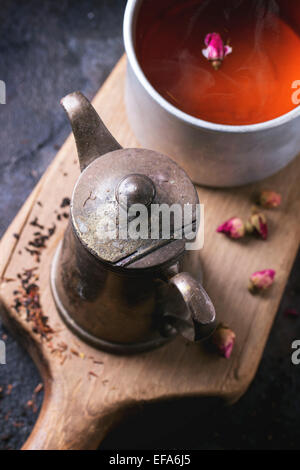 Vintage teapot and aluminum mug of tea, served on wooden cutting board with dry rose buds over dark background. Top view Stock Photo