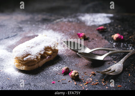 Eclairs with sugar powder, served with dry tea rose buds and vintage cutlery over dark background Stock Photo