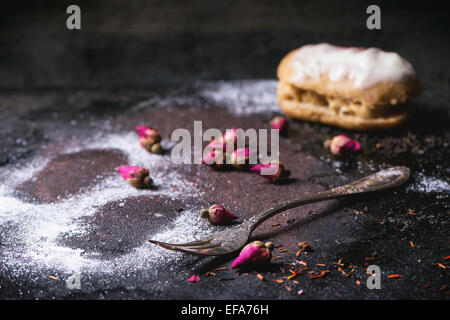 Eclairs and trace of the eclair in sugar powder, with dry tea rose buds and vintage cutlery over dark background Stock Photo