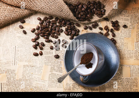 Blue ceramic cup with ground coffee and roasted coffe beans over old newspaper. Top view.
