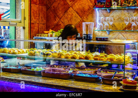 Tapas bar counter in Benidorm, Spain. Glass display case with rows of bocadillos and montaditos sandwiches, salads, toppings. Woman behind counter Stock Photo