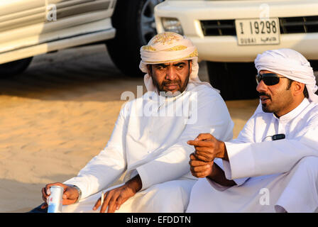 Two men dressed in Arabian clothing sit on sand in front of their 4x4 off-road vehicles. Stock Photo