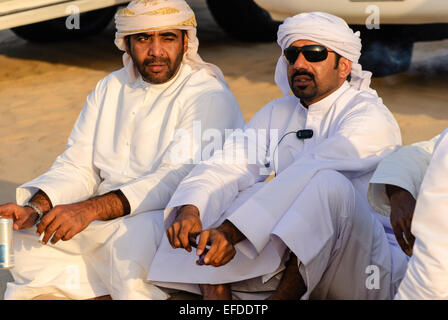 Two men dressed in Arabian clothing sit on sand in front of their 4x4 off-road vehicles. Stock Photo