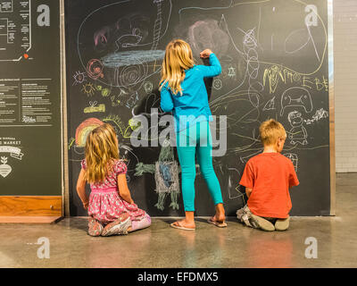 Three young siblings ages 5-10 years old draw with colored chalk on a blackboard in the Santa Barbara Public Market. Stock Photo