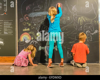 Three young siblings ages 5-10 years old draw with colored chalk on a blackboard in the Santa Barbara Public Market. Stock Photo