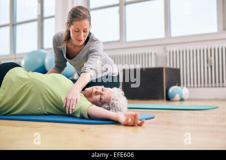Senior women lying on exercise mat doing stretching workout for back muscles with coach assistance. Female trainer helping elder Stock Photo