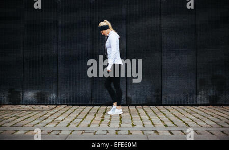 Side view of fitness woman preparing for a city run. Young caucasian woman walking on pavement looking down. Stock Photo